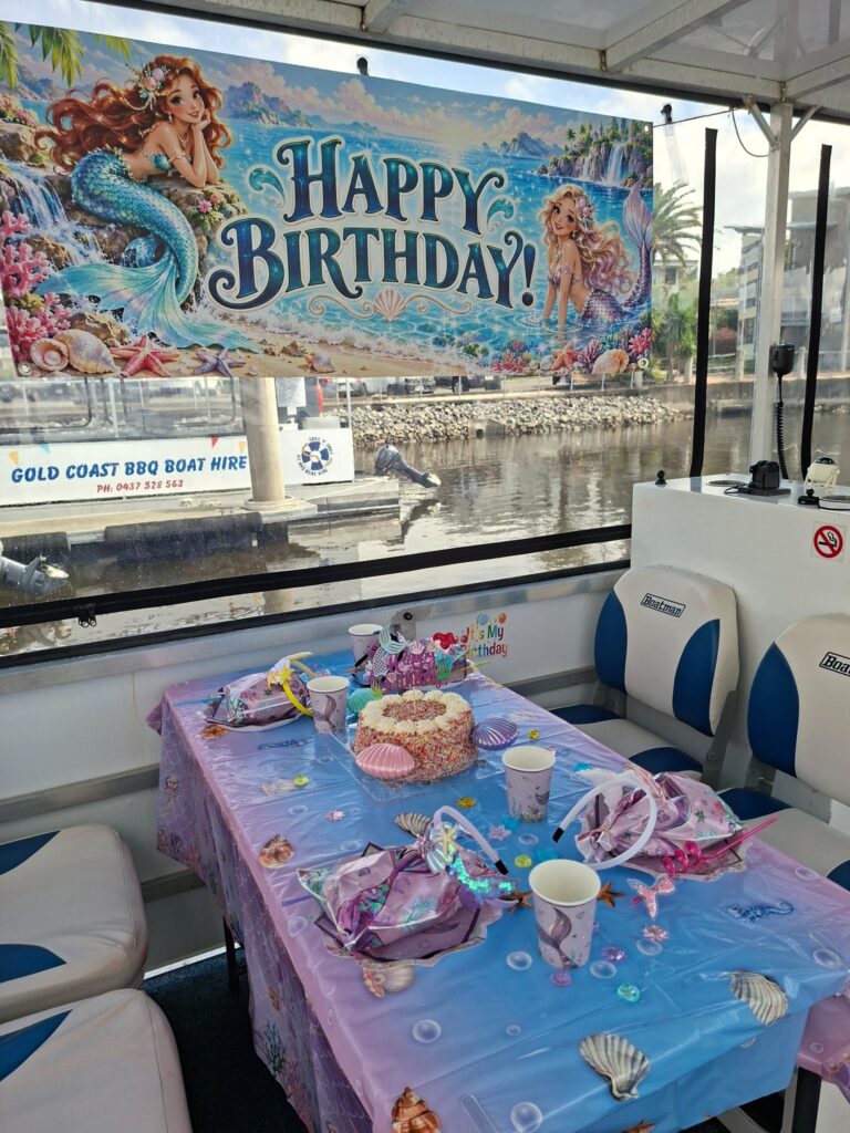 Mermaid birthday party table with cake, headbands, party bags and colourful birthday banner on a BBQ boat